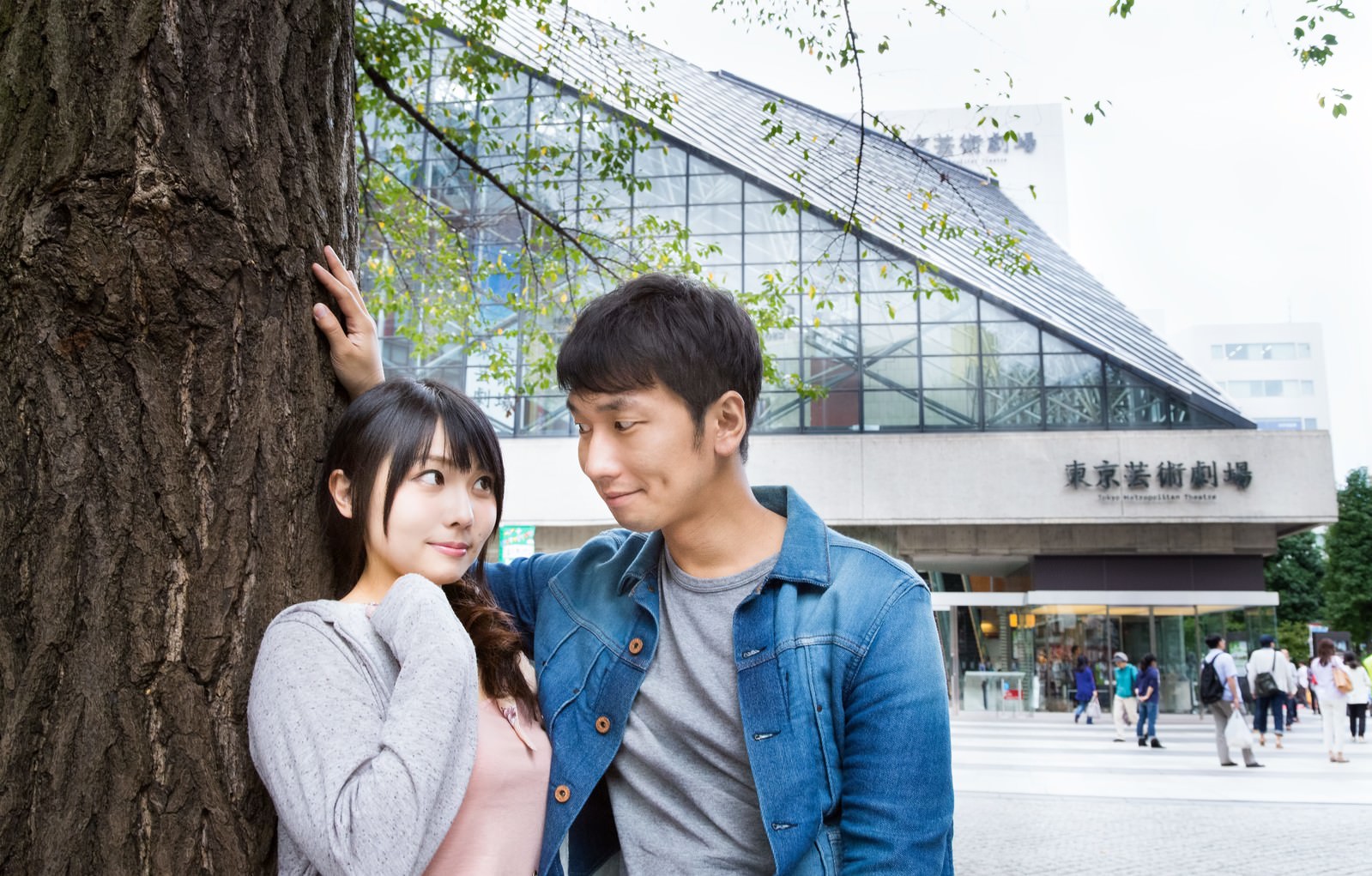 駅前の大きな樹の前で、デニムジャケット姿の男性が樹幹に手をつけて女性に接近するカップル［モデル：大川竜弥 Lala］