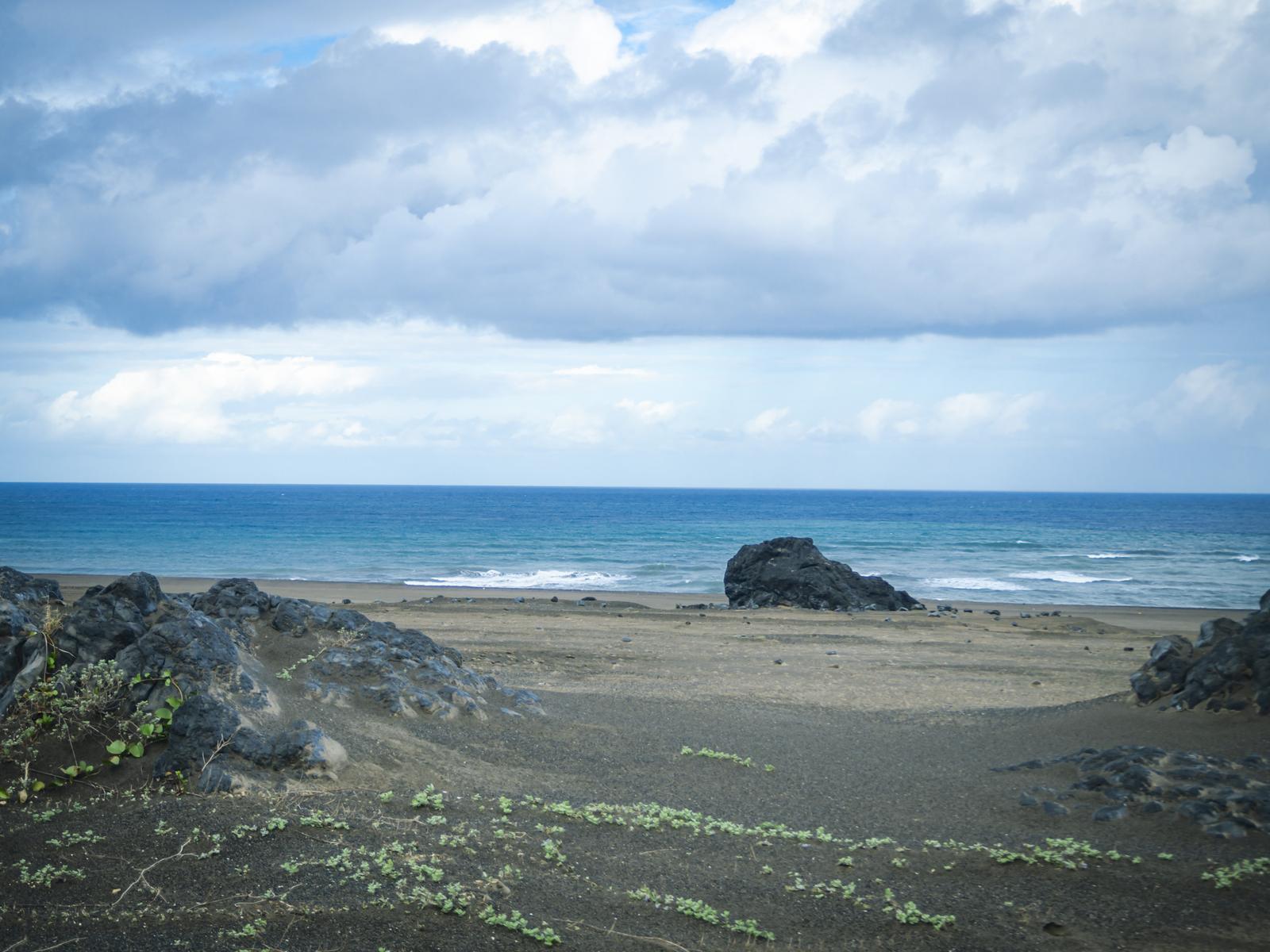 黒い岩が点在する砂浜と青い海、遠くに見える岩礁と白い積雲の空