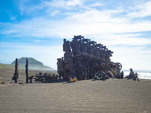 千鳥ヶ浜の沈船のエンジンなどの残骸と強い風で流れるしぶきに霞む砂浜の奥に立つ摺鉢山