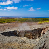 青空と白い雲が広がる鶯地獄の噴気孔と沖に見える監獄岩の写真