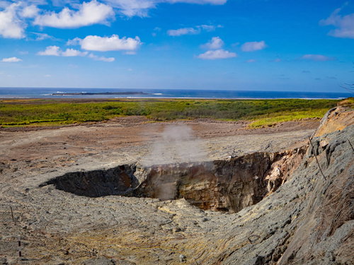 青空と白い雲が広がる鶯地獄の噴気孔と沖に見える監獄岩