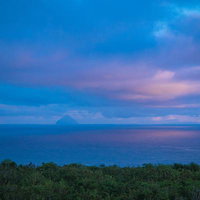 夕焼け色の空と同じ色に染まる海の先にかすむ南硫黄島の写真