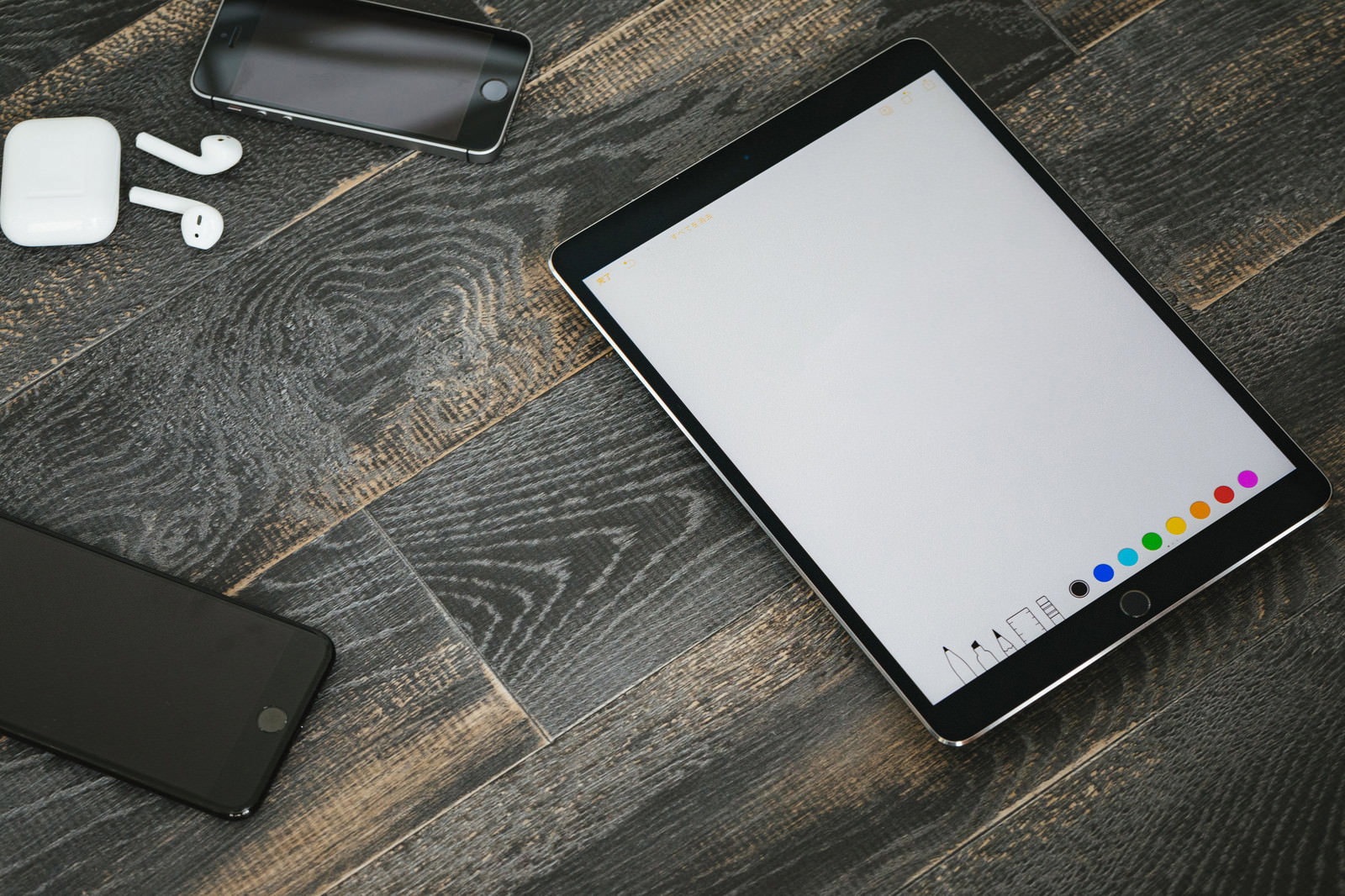 木目テーブルの上に置かれたiPadとスマートフォン、AirPodsを俯瞰で撮影