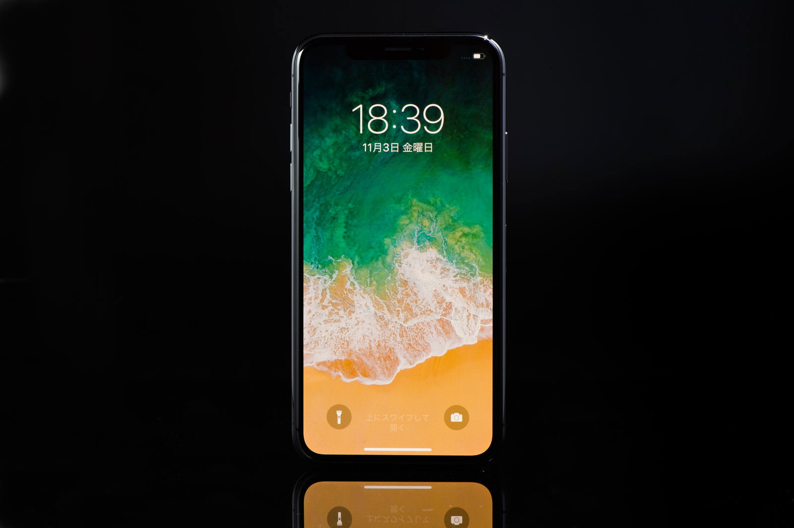 黒い背景に置かれた画面点灯中のiPhone X、ロック画面に海岸の風景表示