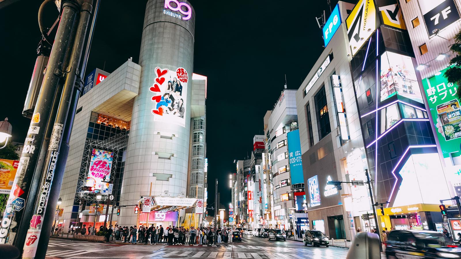 Wide-angle night shot of SHIBUYA 109 tower on the left with a red heart ad, neon-lit buildings on the right, pedestrians near a crosswalk.
