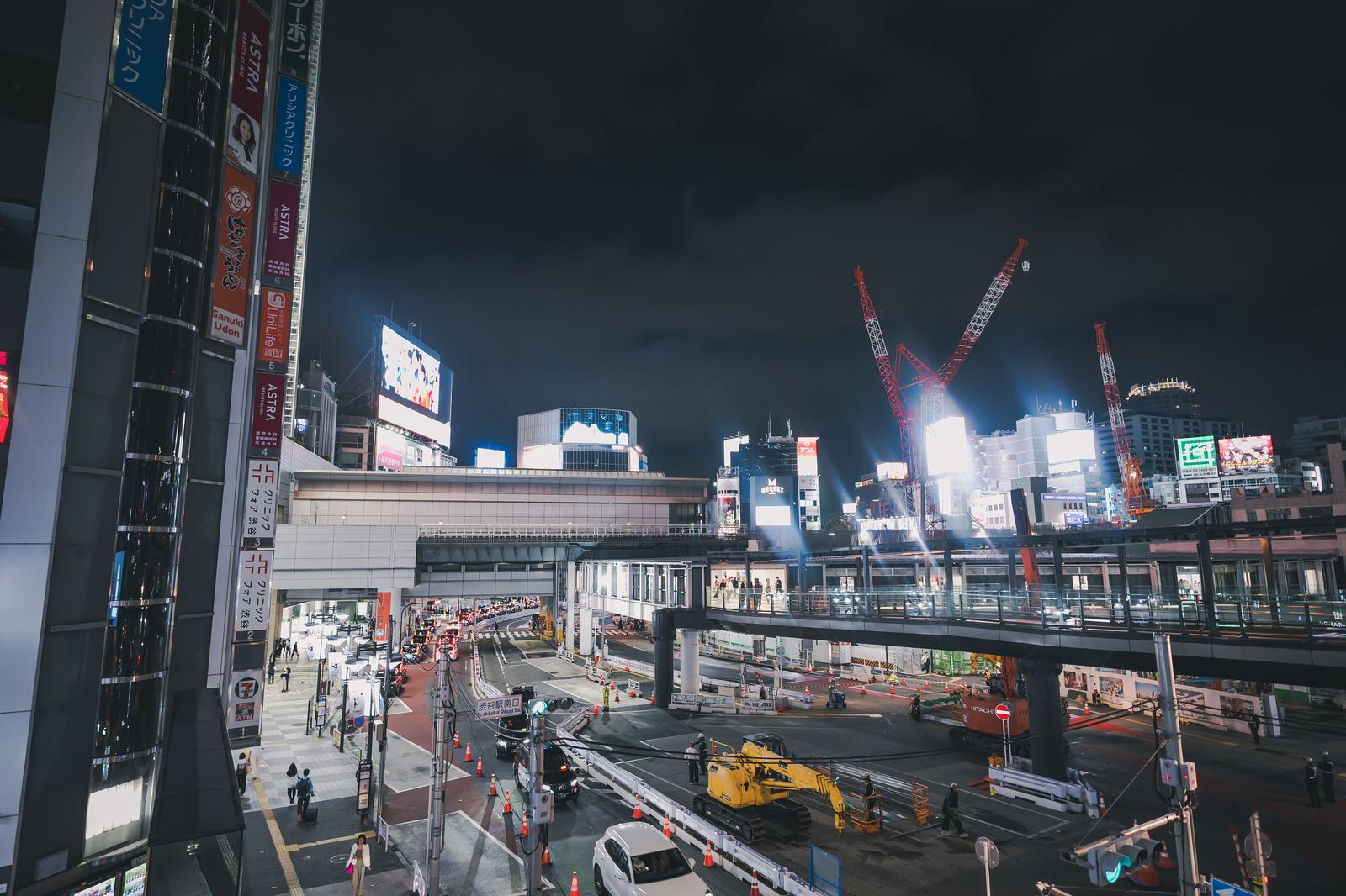 夜間の渋谷駅南口で、クレーンと点灯したネオン看板に照らされた工事現場と人通りのある光景