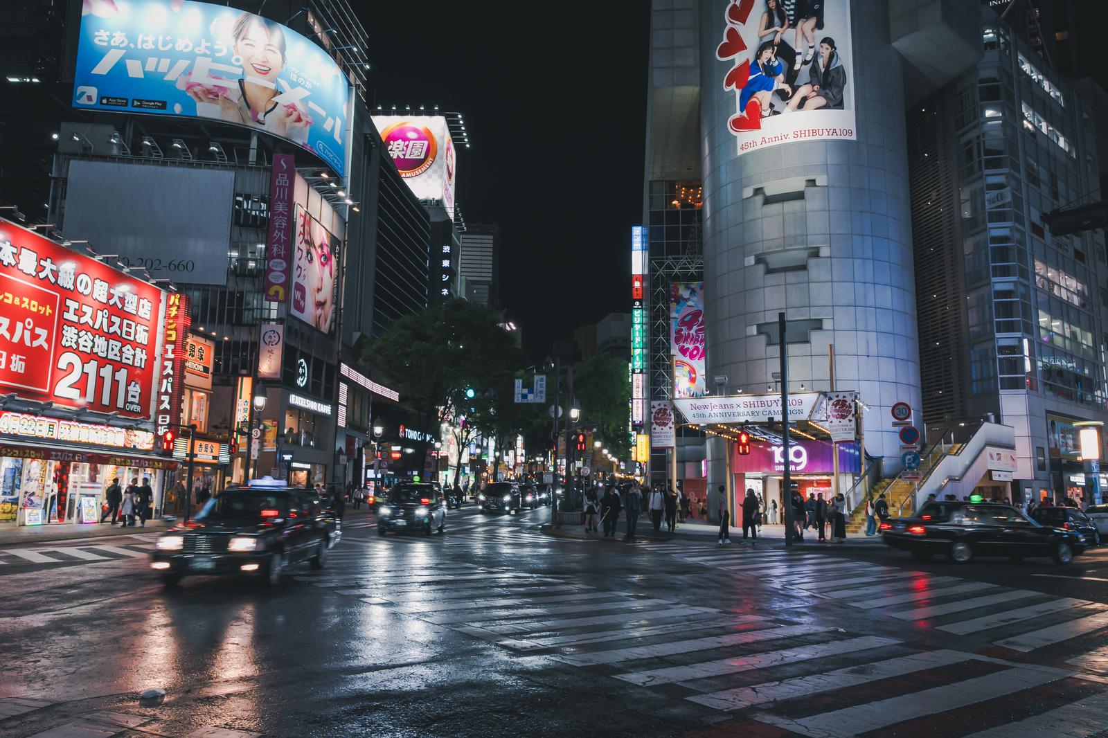 Night view of Shibuya 109 crosswalk on Dogenzaka, Tokyo, after rain. Wet road reflects neon signs and tail lights; pedestrians and taxis visible.