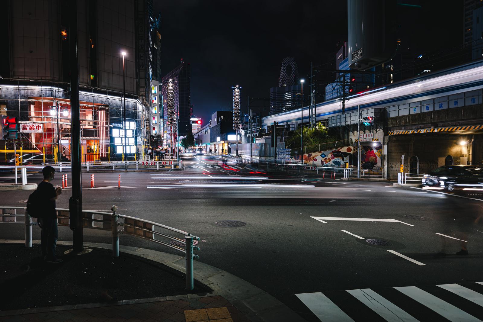 新宿の交差点を横断する人々と通過する電車の光跡が見える夜間の街並み