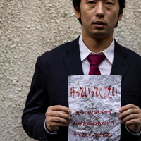 濃紺スーツの男性が赤い文字の怪文書を両手で掲げるポートレートの写真