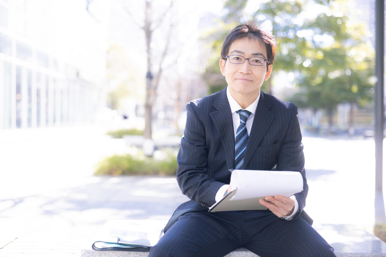 街中で資料を持ち微笑む眼鏡をかけたスーツ姿の営業部長［モデル：ガーミー］