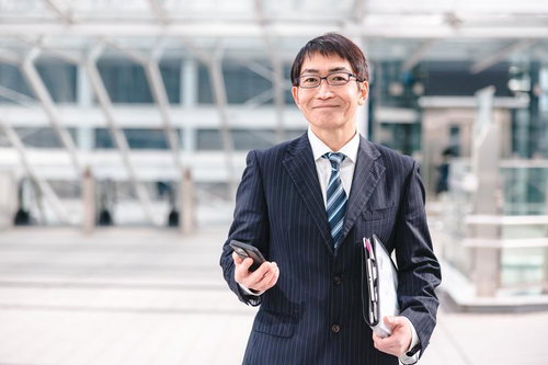 ニコニコしたアラフィフ部長がスマートフォンを片手に携帯電話を持ちながら立つ