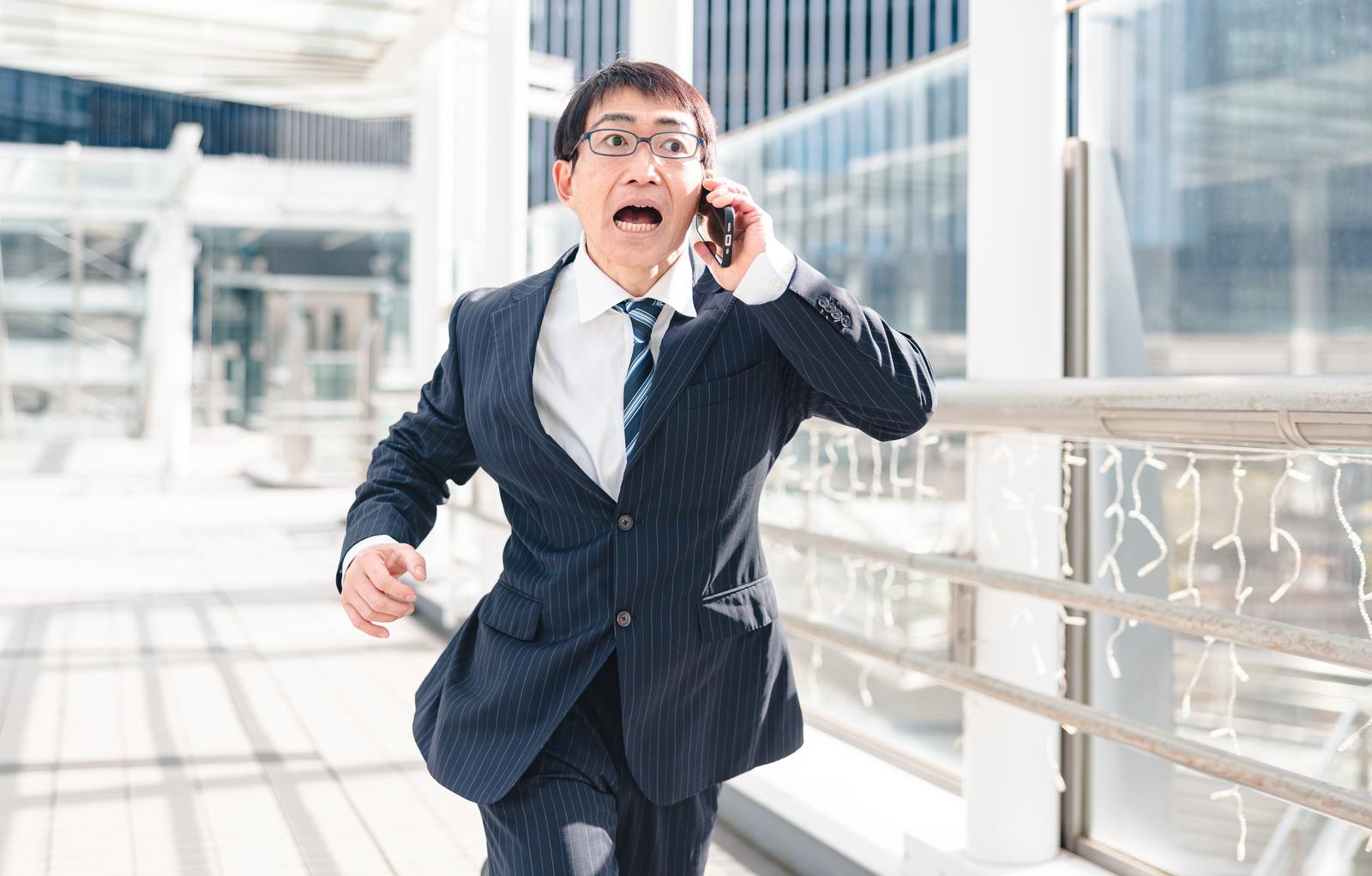眼鏡をかけた男性がスマホを持ちながら走っており、高層ビルが背景に見える［モデル：ガーミー］