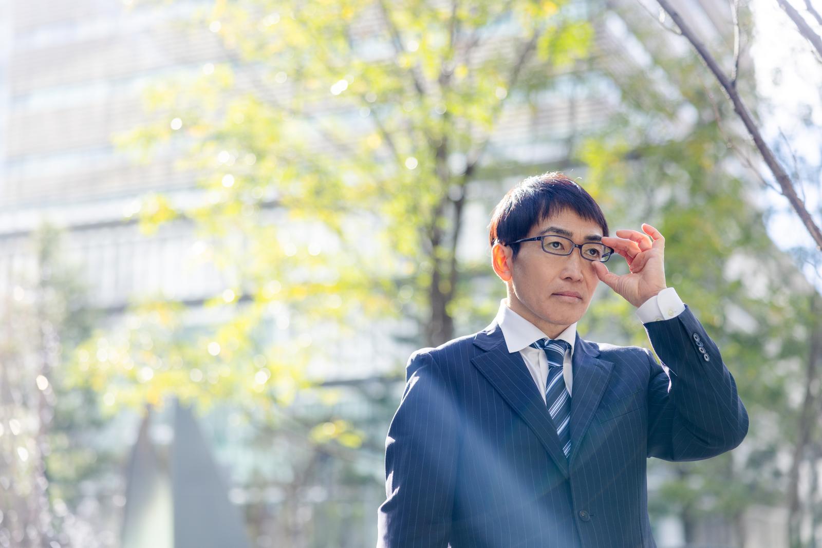 メガネを持ち上げるスーツ姿の営業男性。新緑の街路樹が背景［モデル：ガーミー］