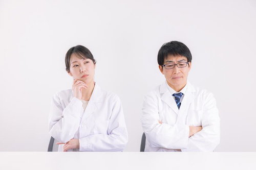 腕を組んで険しい顔をする白衣の男性医師と悩む表情の女性研究者
