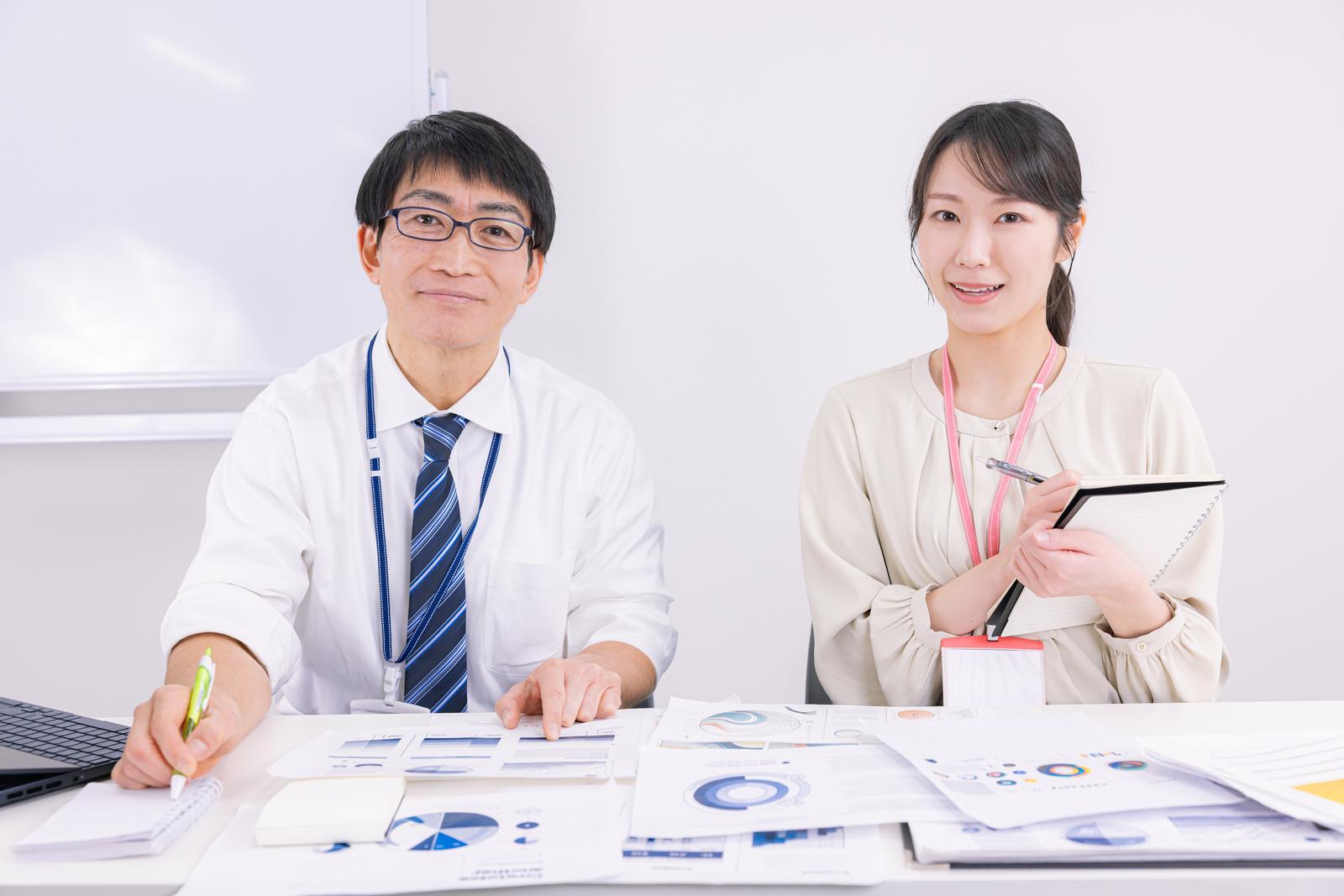 資料を指しながら説明する眼鏡の男性と、メモを取る女性が机に向かっている［モデル：ガーミー SAKI］