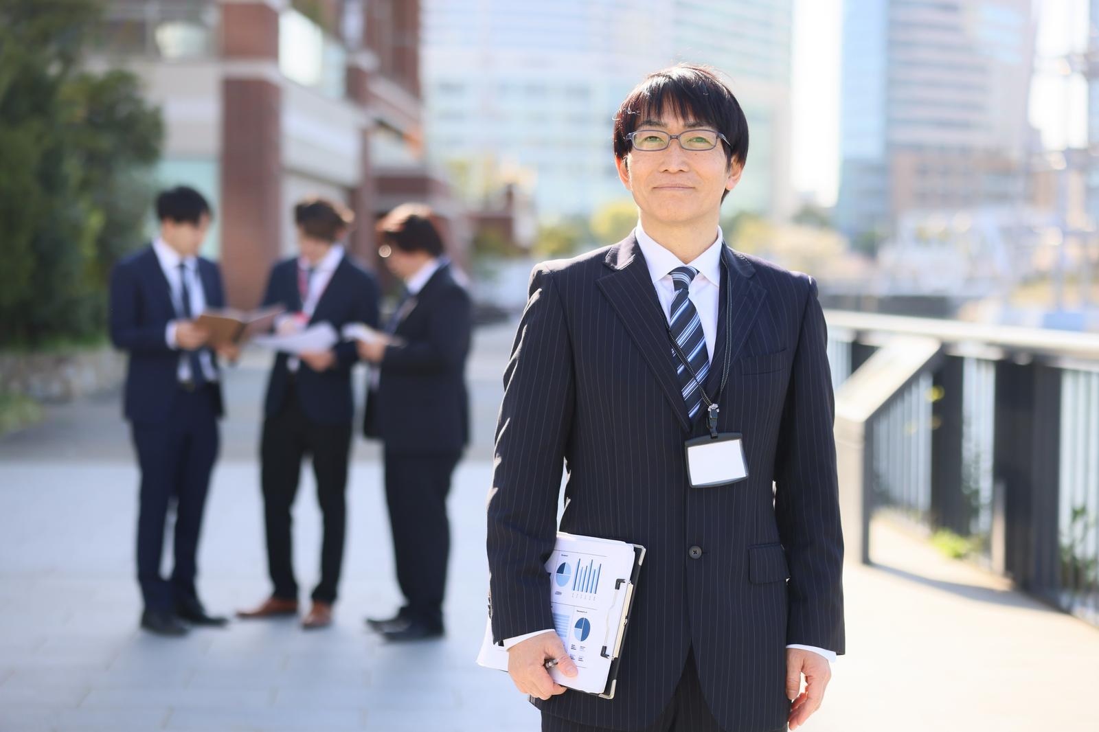 スーツを着た眼鏡の男性上司が書類を持ってカメラを向き、背景に同僚たちとビル群が見える［モデル：ガーミー］