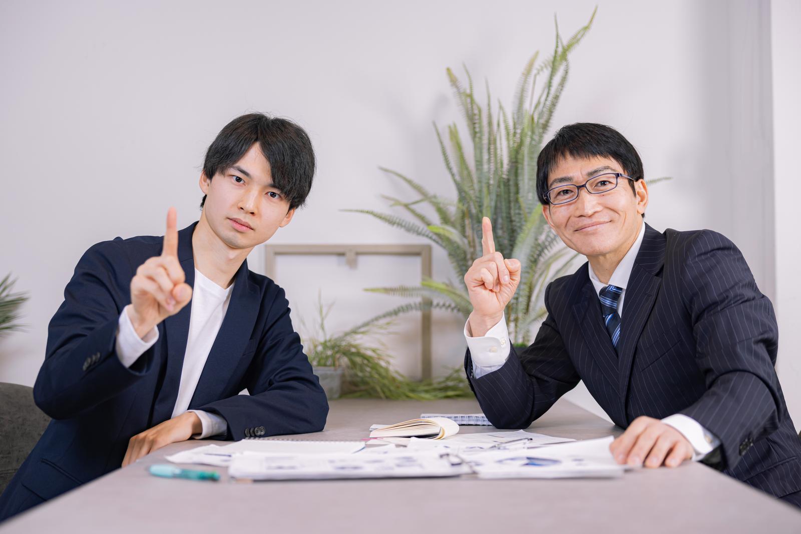 机の上に資料を置いて、指を立てるジェスチャーをする2人の会社員［モデル：たくみ ガーミー］