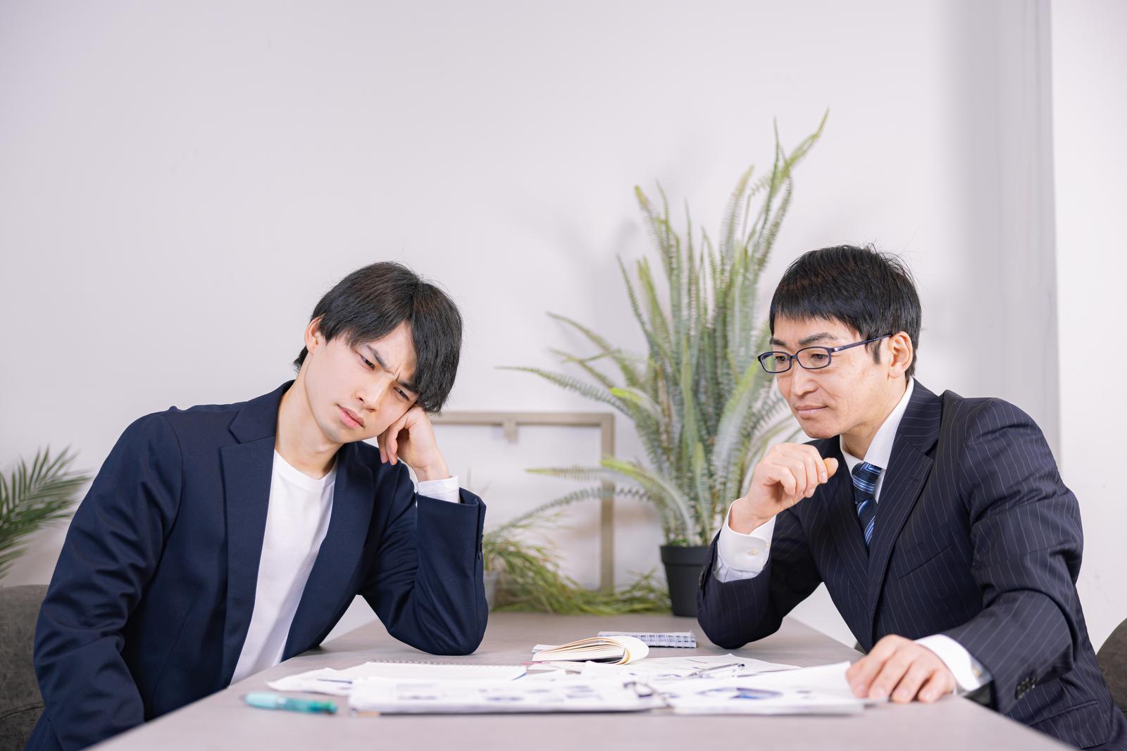 資料を前に考え込む紺色のスーツを着た2人のビジネスマン［モデル：たくみ ガーミー］