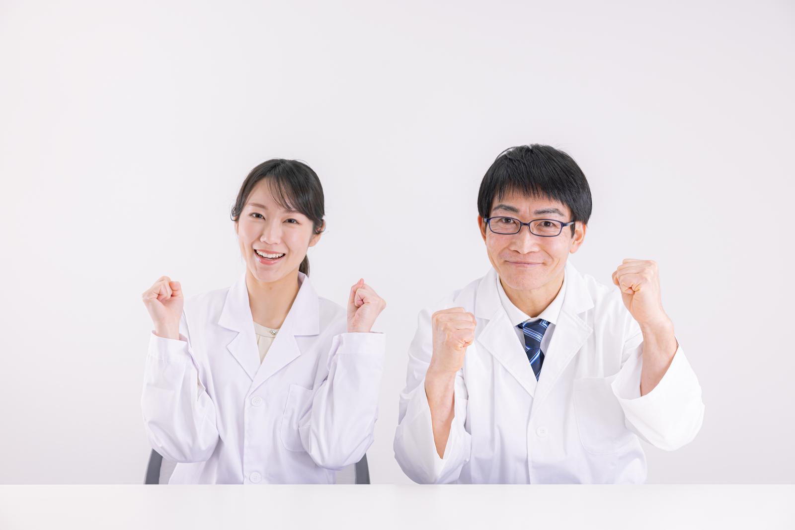白衣を着た医師の男性と女性がガッツポーズをしながら笑顔で立つ［モデル：ガーミー SAKI］