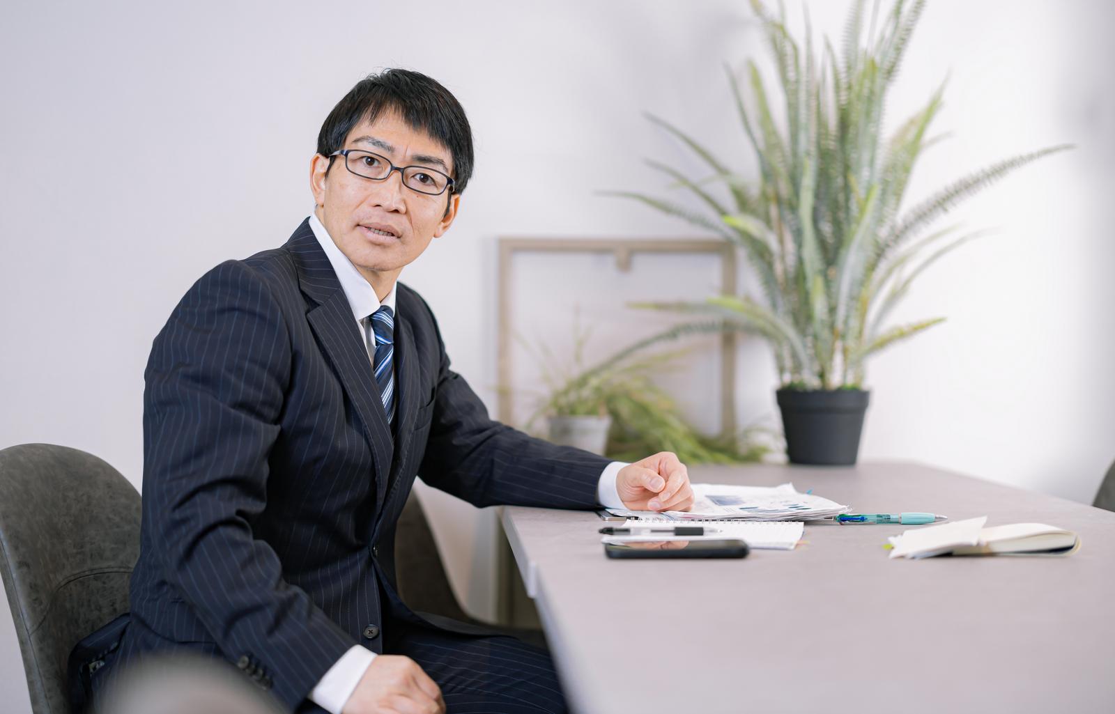 オフィスの机で書類を前に激怒し、左を向いて話すスーツ姿の眼鏡の男性［モデル：ガーミー］