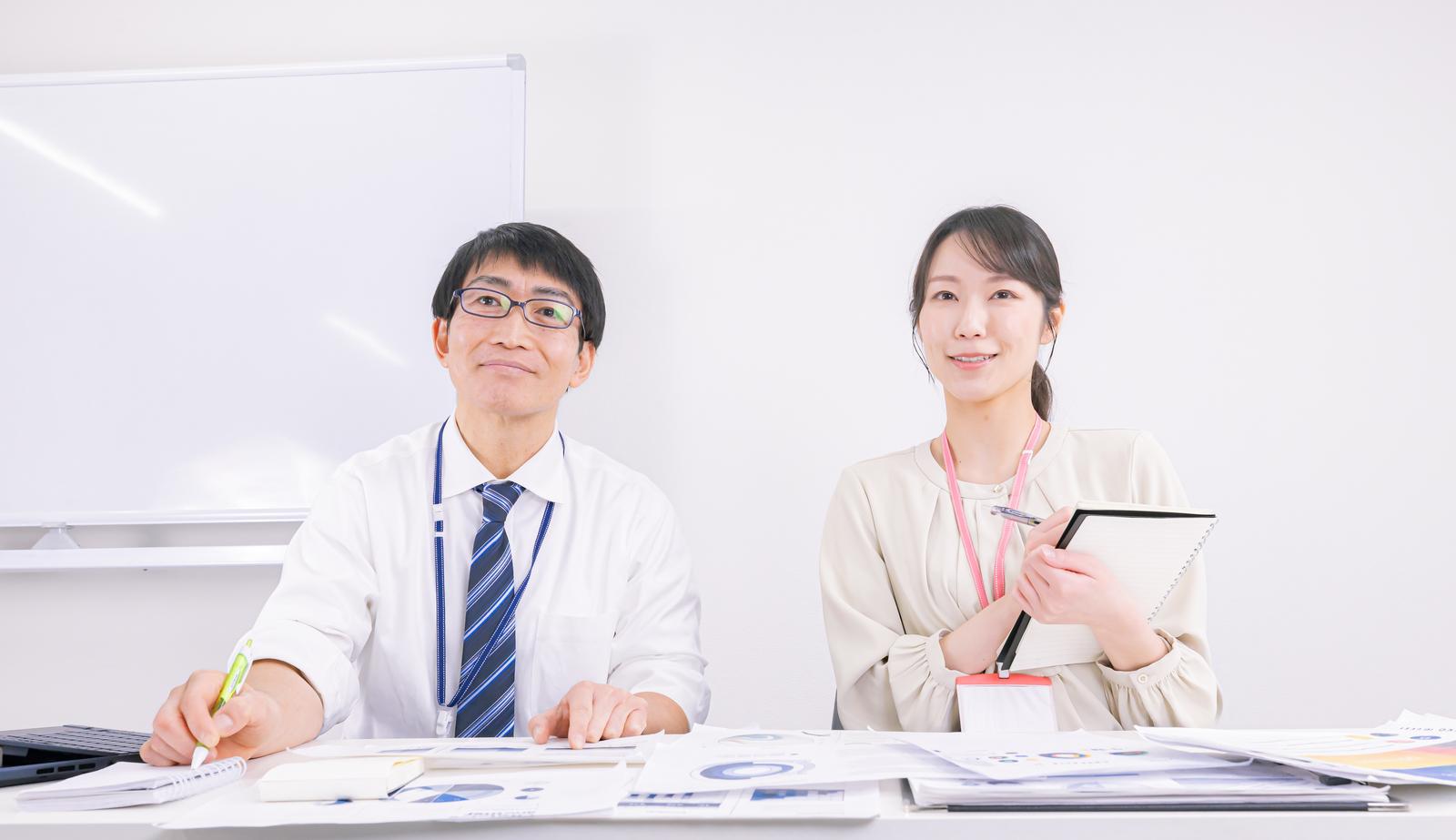 ホワイトボードを背景に資料を確認しながら会議をする男性と女性の職員［モデル：ガーミー SAKI］