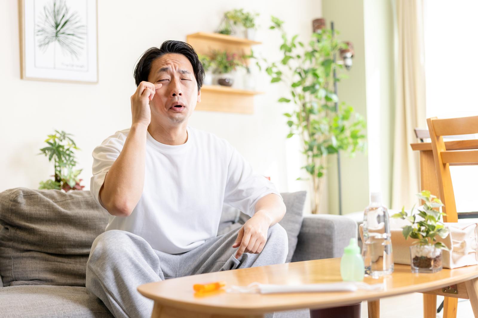 ソファーに座り目元を押さえて苦しそうな表情をする男性［モデル：大川竜弥］