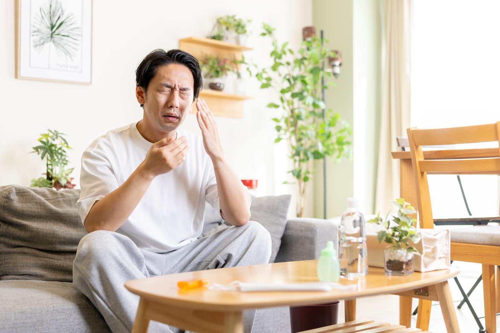 ソファーに座り鼻をかむ表情の苦しい男性［モデル：大川竜弥］