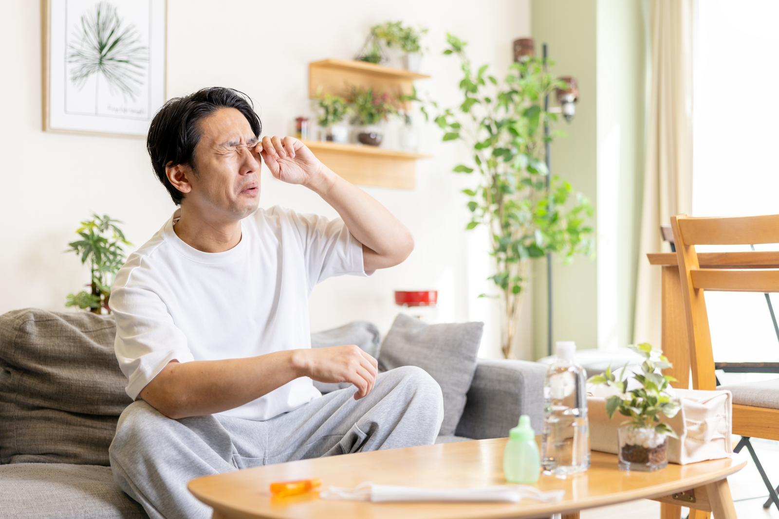 花粉症で目をこすり困った表情の男性がソファーに座っている［モデル：大川竜弥］