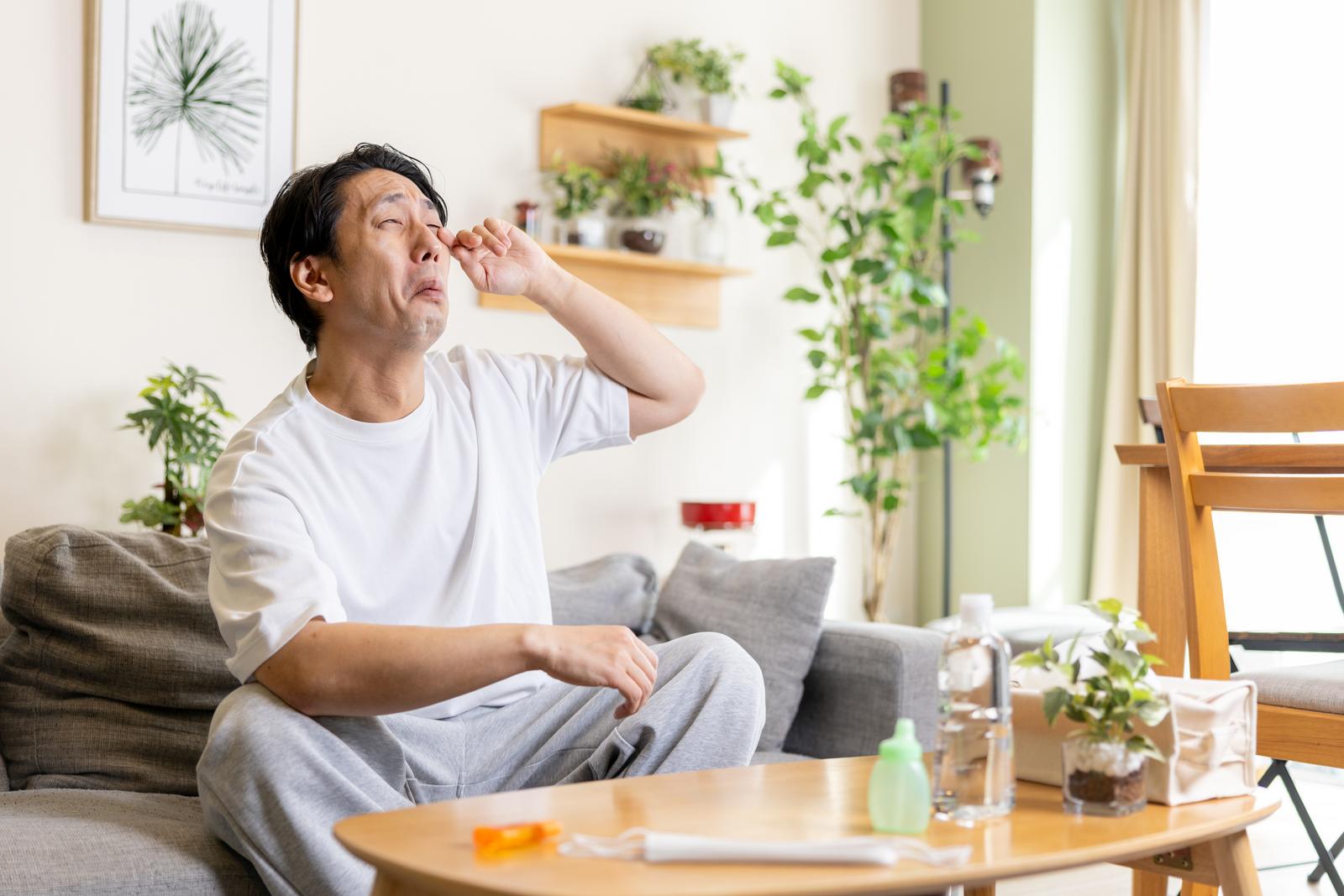 花粉症で目のかゆみをこする男性がソファーに座っている［モデル：大川竜弥］