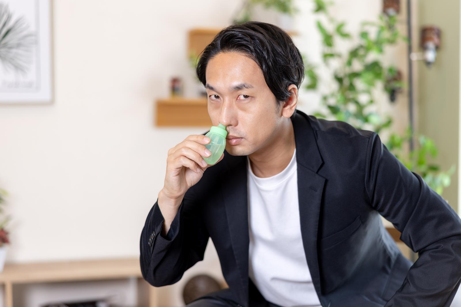緑色の鼻洗浄器を使って鼻うがいをするビジネスジャケット姿の男性［モデル：大川竜弥］