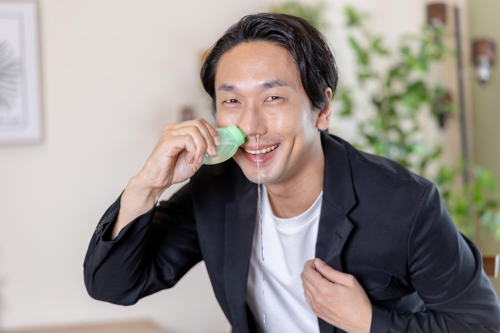 グリーンの鼻洗浄容器を使いながら笑顔で鼻うがいする、黒いジャケット姿の男性［モデル：大川竜弥］