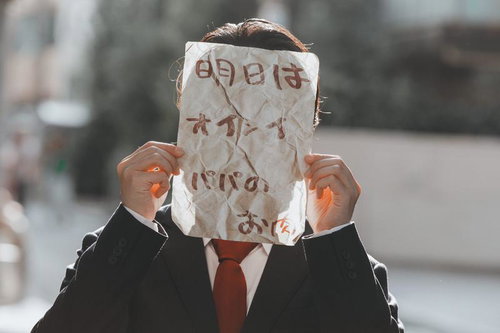 紙袋で顔を隠した黒スーツの男性が怪文書を掲げるポーズ