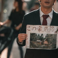 「この家危険」の怪文書を持つスーツ姿の男性の写真