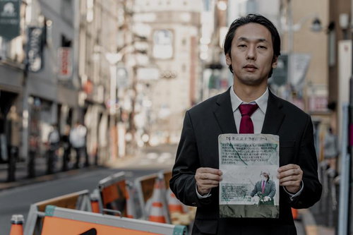 赤ネクタイのスーツ姿で怪文書を掲げる男性