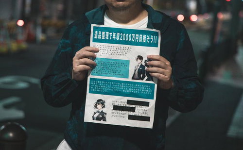 夜の街角で高収入勧誘の怪しいチラシを持つ黒いパーカーの人物