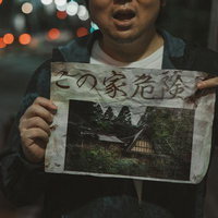 夜の街頭で「この家危険」と書かれた古い怪文書を指差す男性の写真
