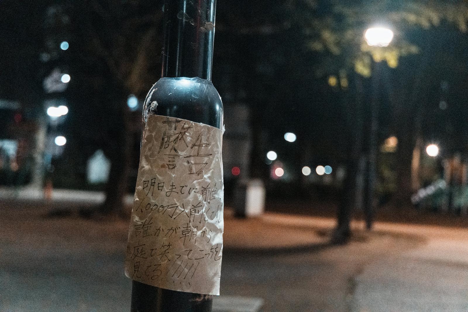 Crumpled handwritten warning note in Japanese script taped to a dark metal street bollard at night, with blurred bokeh city lights in the background.
