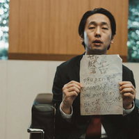 応接室で手書きの怪文書を掲げる黒いスーツの男性の写真