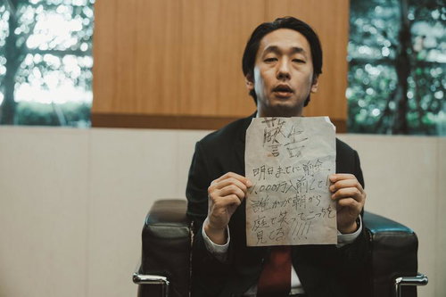 応接室で手書きの怪文書を掲げる黒いスーツの男性