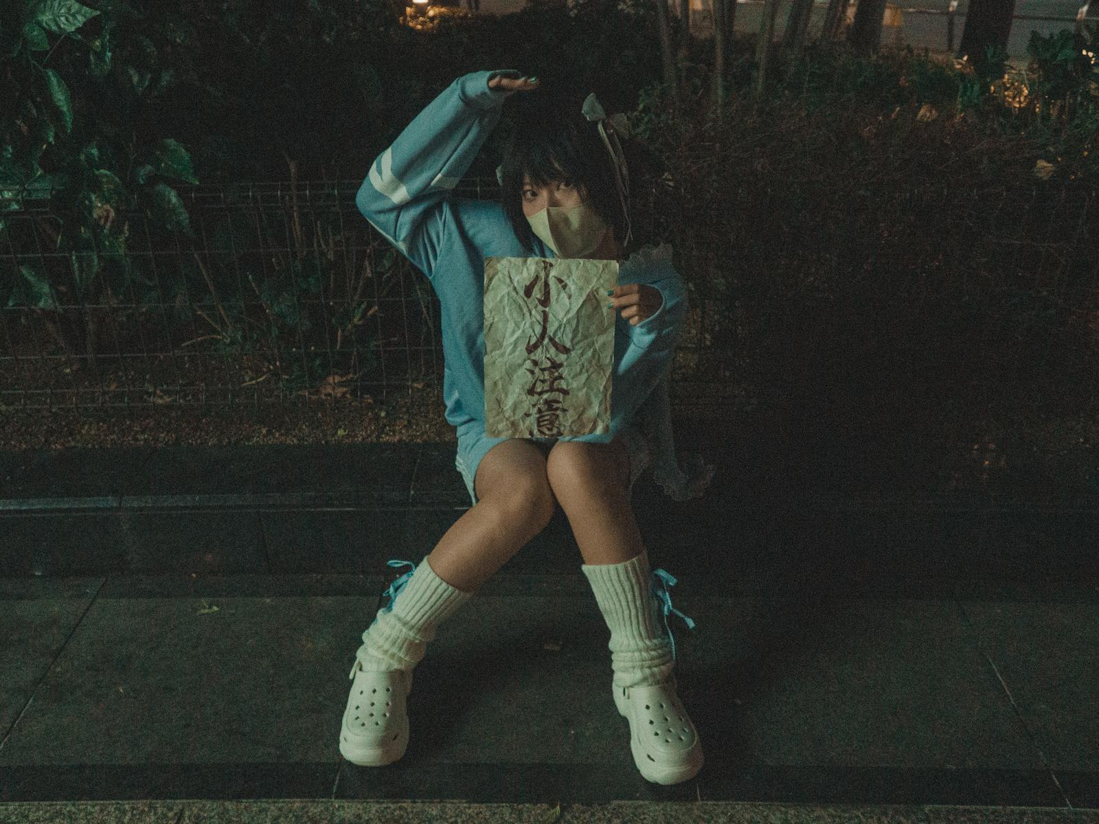 夜の路上で青い服を着た女性が怪文書らしき紙を手に持って読んでいる［モデル：緋真煉］