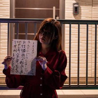 住宅街の手すりに座り怪文書を掲げる赤い服の女性の写真