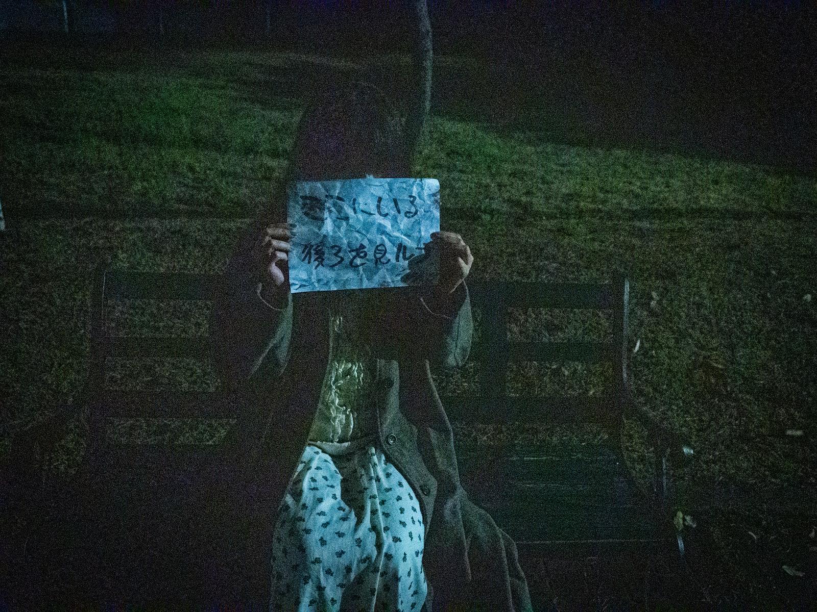 夜のベンチで青い紙に書かれた怪文書を両手で掲げる女性［モデル：ラミエル］