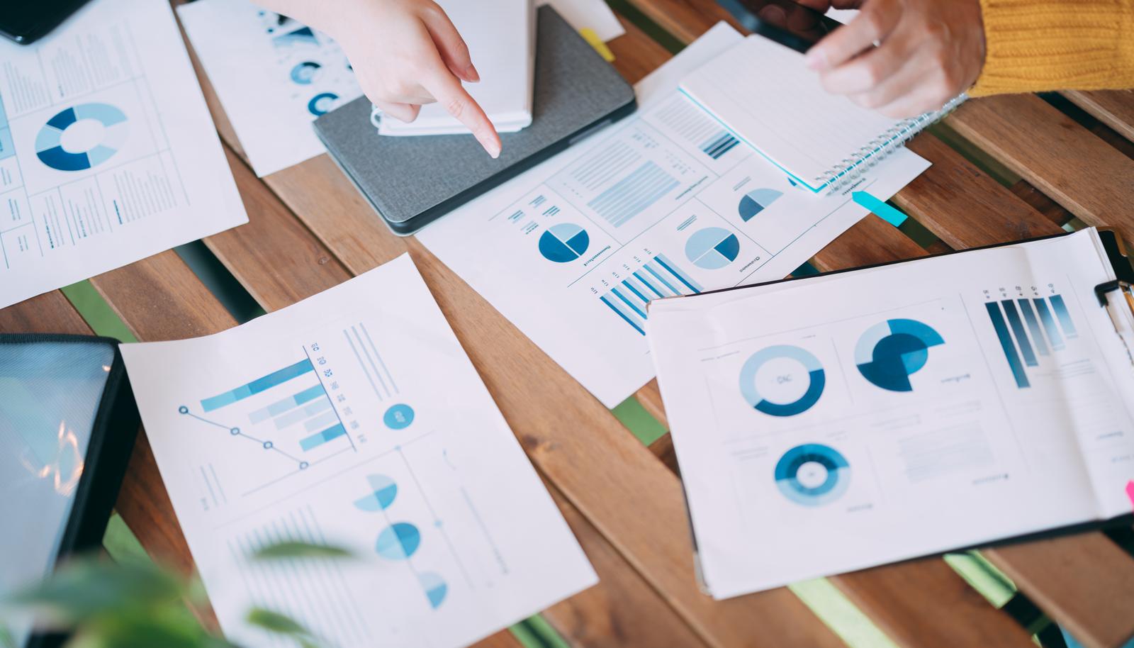 木製テーブルの上に複数の資料とグラフが散らばり、男性がタブレットを操作しながら書類を指差している会議シーン