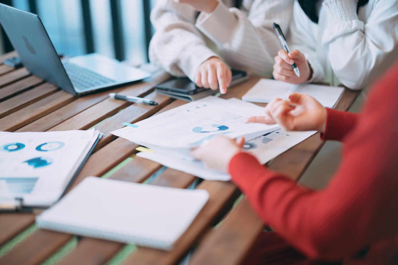 グラフ資料を見ながら打ち合わせする複数人の手が机の上に置かれている