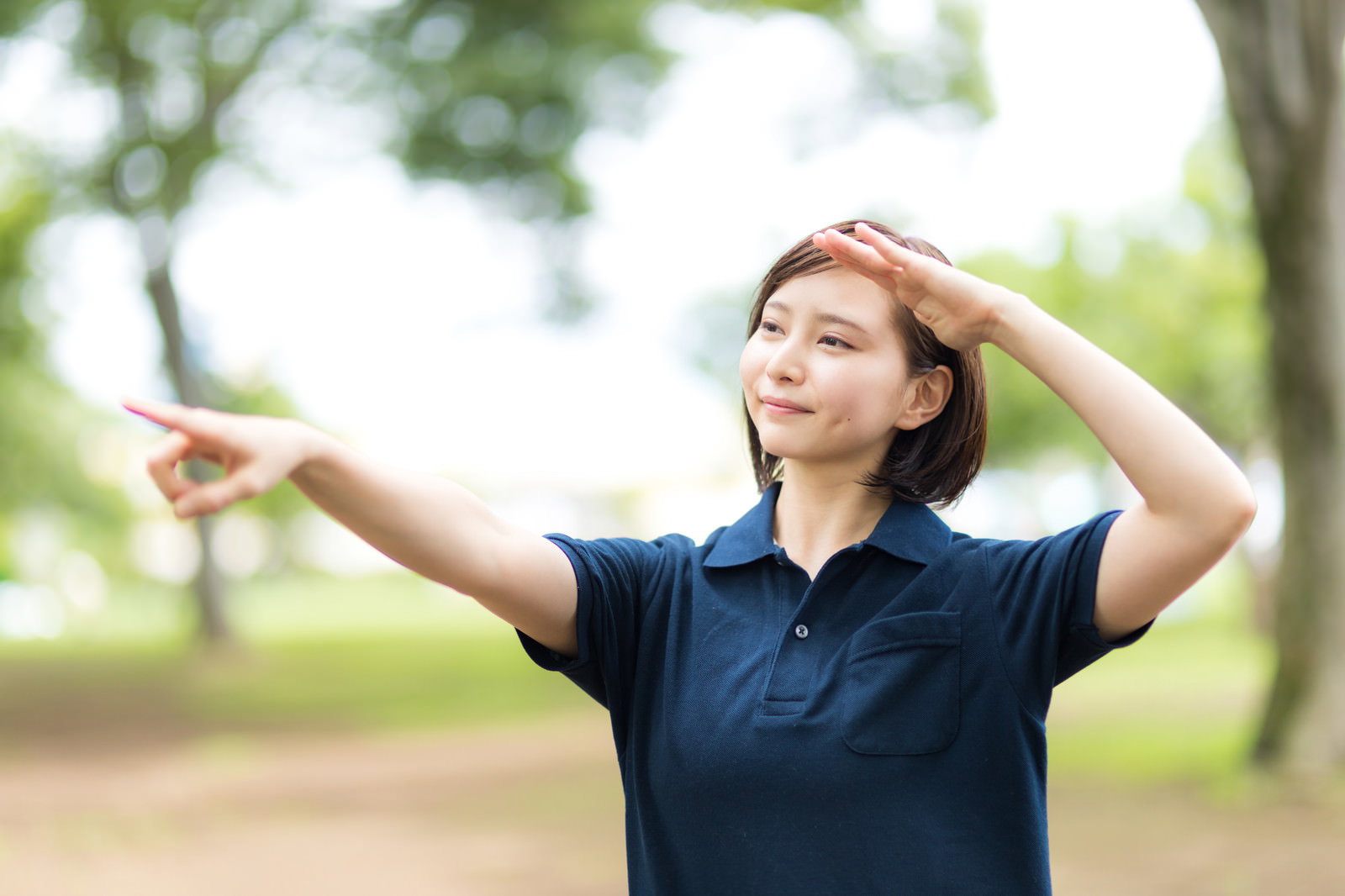 紺色の制服を着た女性介護士が片手を額にかざし、もう一方の手で日陰を指差している［モデル：yumiko］