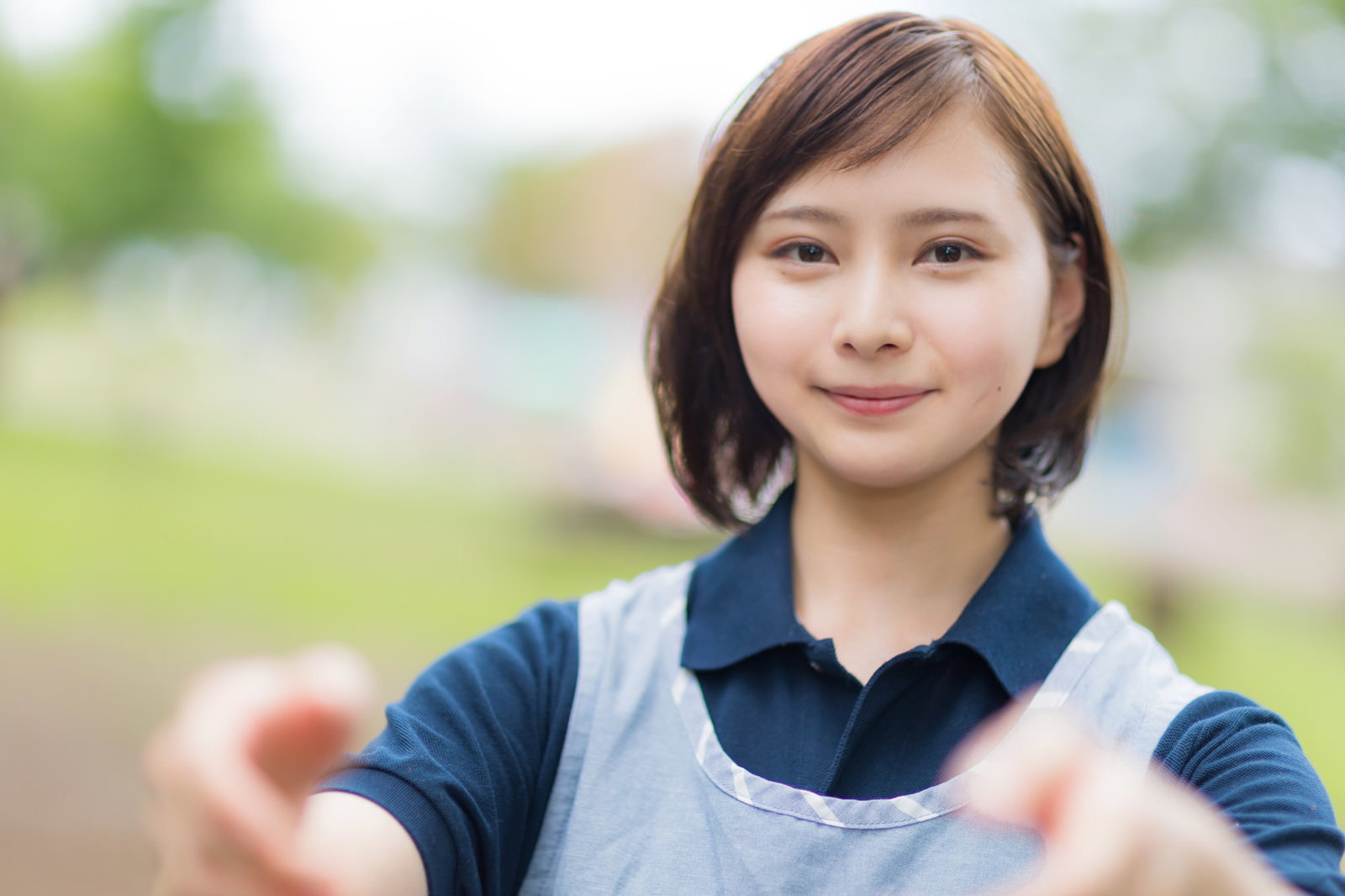 青いエプロンを着た介護士の女性が笑顔で両手を差し伸べている様子［モデル：yumiko］