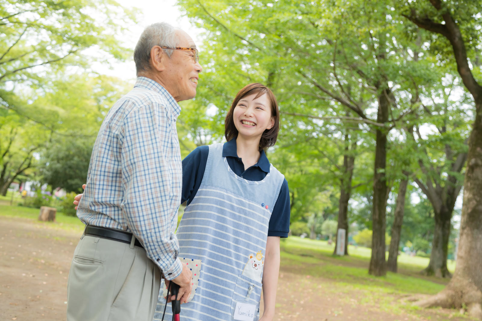 緑豊かな公園で高齢者の男性と並んで散歩する介護士の女性［モデル：まーちゃん yumiko］