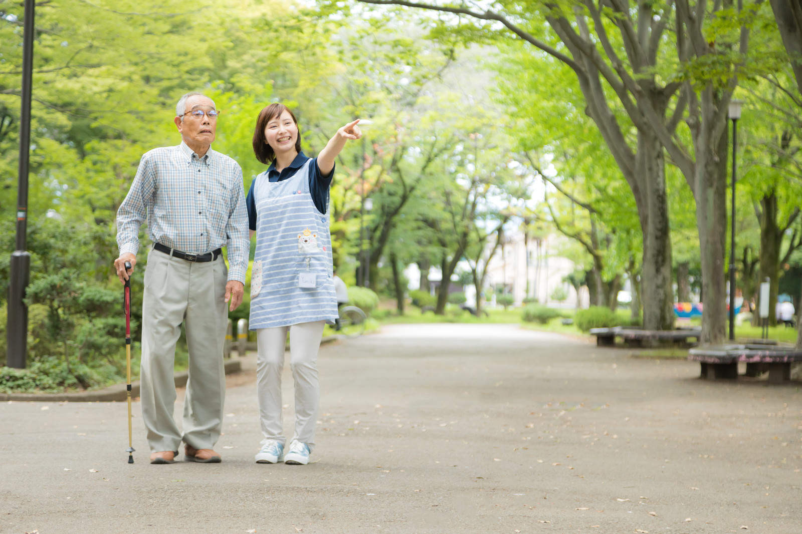 新緑の公園で腕を組んで散歩する高齢男性と介護士の女性［モデル：まーちゃん yumiko］