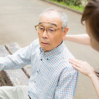疲れてベンチに座る老人に声をかける女性介護者の写真