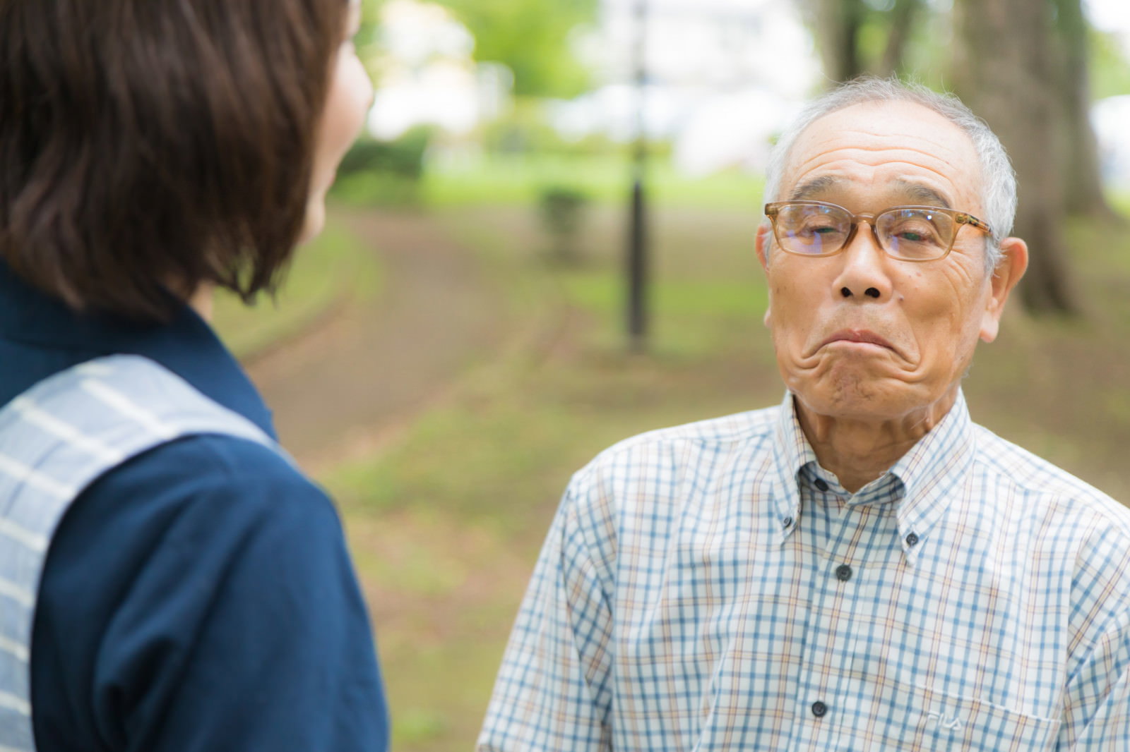公園で口をへの字にした眼鏡の高齢男性と話しかける女性ヘルパー［モデル：まーちゃん yumiko］